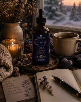 A dark amber dropper bottle labeled 'Courage in the Dark' stands on a wooden surface by a window, surrounded by dried flowers in a vase, a lit candle, a ceramic mug, a notebook, and a pen. A snowy outdoor scene is visible in the background.