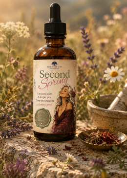 An amber glass dropper bottle labeled 'Second Spring' by The Root Art Apothecary, styled outdoors on a stone surface with wildflowers, dried herbs, and a mortar and pestle in a sunlit meadow.