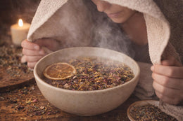 May include: A beige ceramic bowl filled with steaming water and dried herbs, with a slice of dried orange. A person is covered with a towel, inhaling the steam. A lit candle is in the background, creating a relaxing spa-like atmosphere.